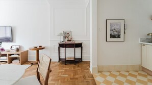 Ein Zimmer mit Holzboden im Fischgrätmuster, einem Esstisch mit Stühlen, einem kleinen Beistelltisch mit Lampe, einem gerahmten Bild an der Wand und einer karierten Fußmatte neben der Tür.