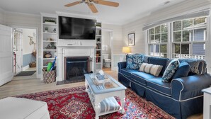 A living room with a fireplace, a television, a blue sofa, and a ceiling fan. - Grace Cottage, St Michaels Walk to everything! (Saint Michaels)