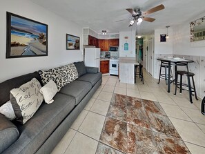A modern living room with a grey sofa, a patterned throw pillow, and a large painting of a beach scene. - Casa Del Mar 394 | Coastal Condo w/ Private Balcony & Ocean Views (Galveston)