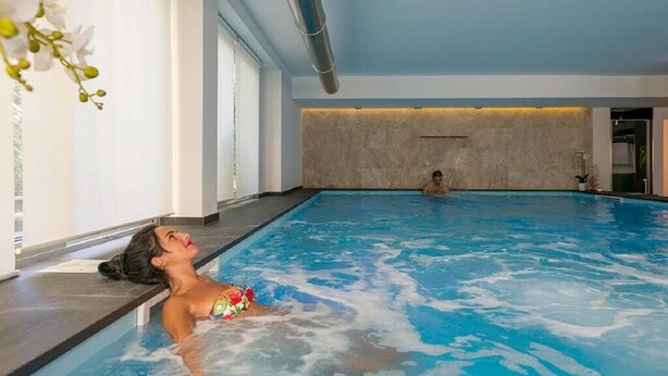 An indoor swimming pool with clear blue water, a person relaxing on the edge, and another person in the background. - Zenit Hotel (Giulianova)