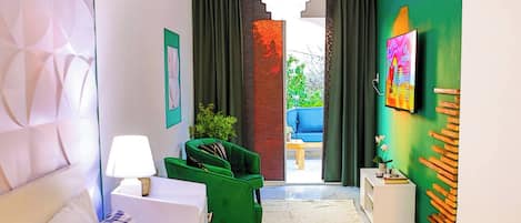 A hotel room with a bed, a green armchair, a white nightstand, a lamp, a TV on the wall, and a view of the outside balcony.