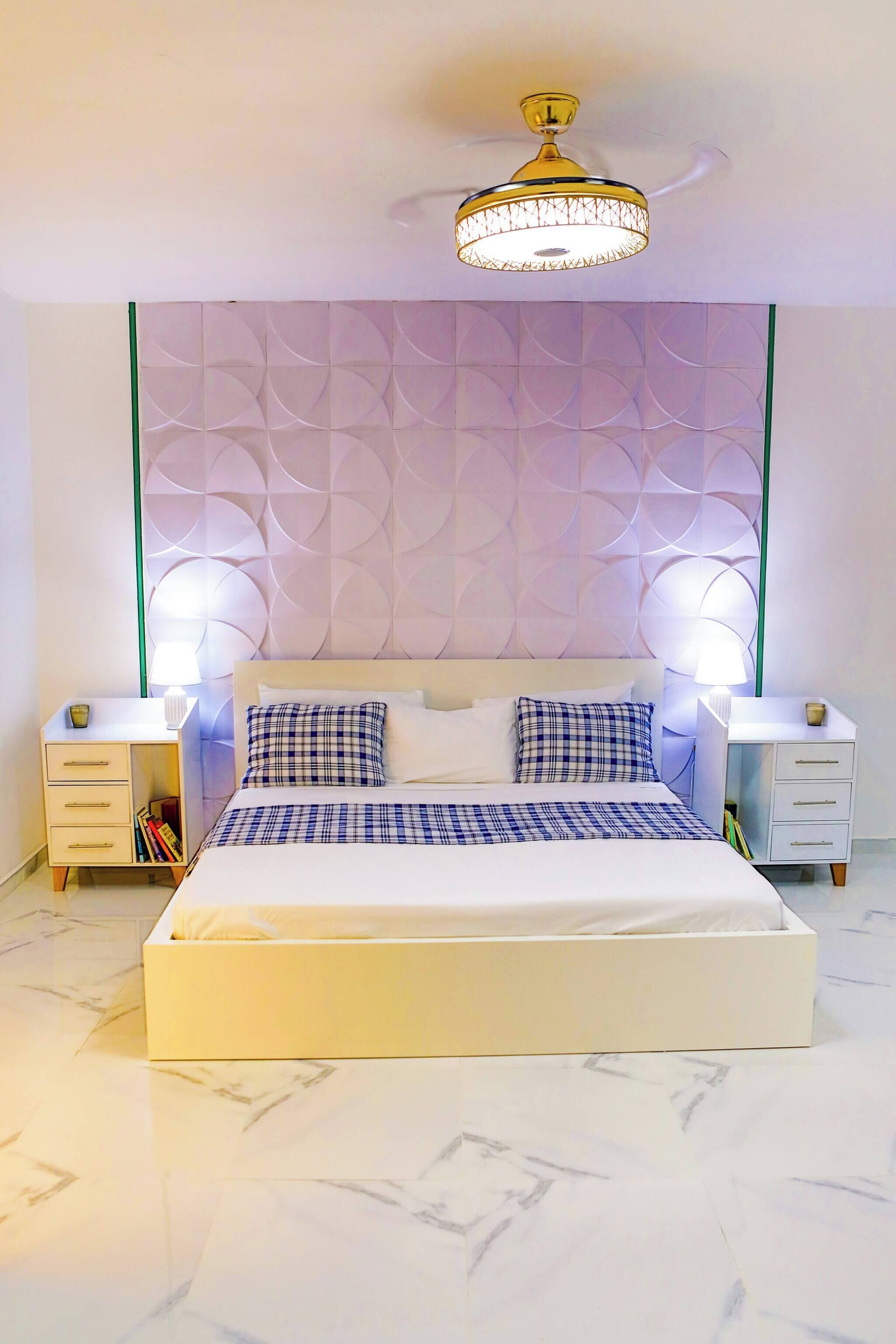 A modern bedroom with a large bed, two bedside tables, a decorative wall panel, and a ceiling light fixture.