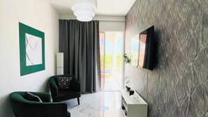 Ein modernes Wohnzimmer mit einer Chandelieber, einem Flachbildfernseher, einem grünen Sofa, einem weißen Konsolentisch und einer Wand mit Muster.