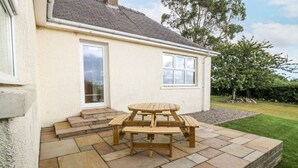 Un patio con mesa y bancos de madera, una casa blanca con techo gris y un césped con árboles.
