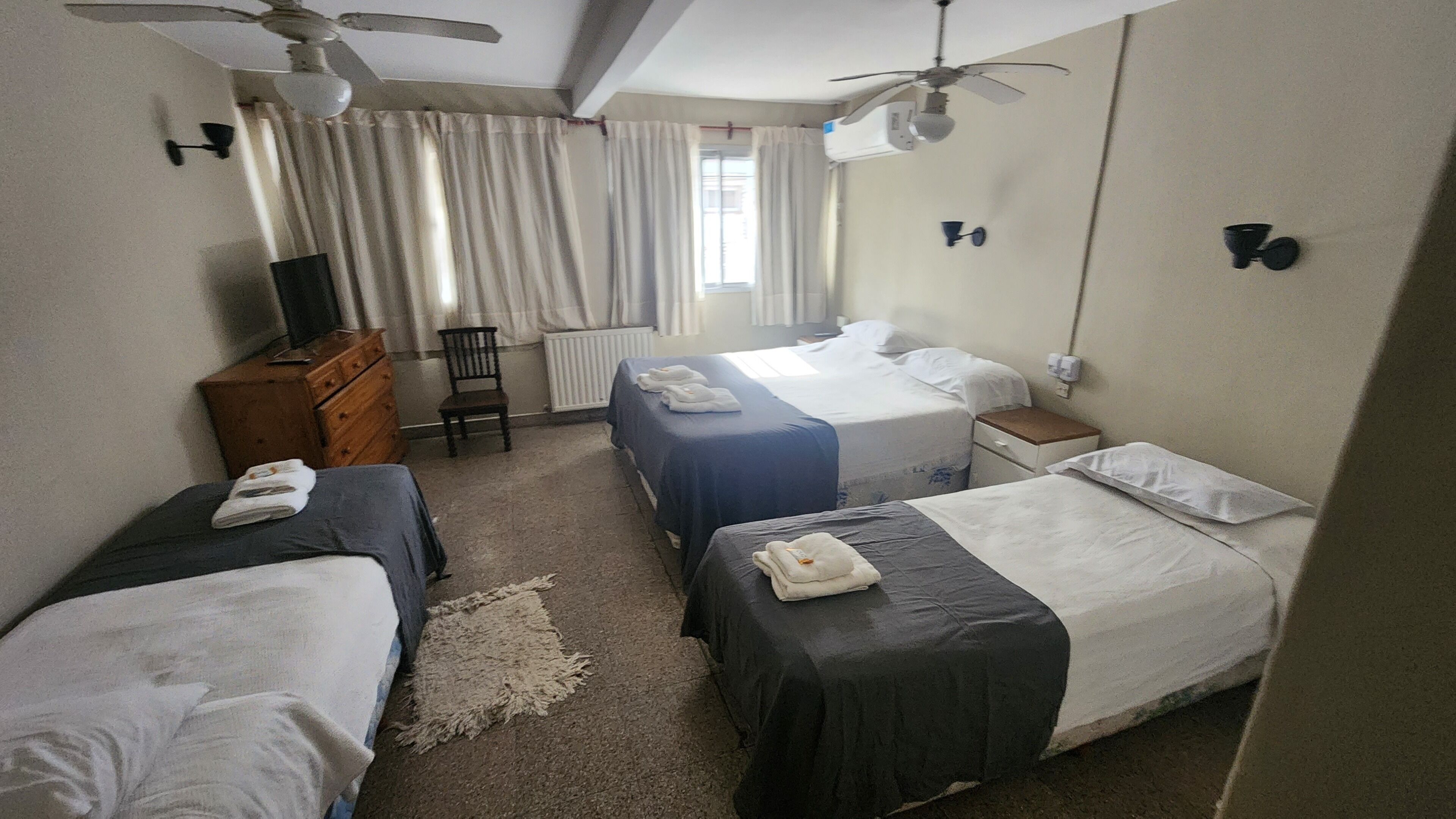 Habitación de hotel con dos camas, un aparador, una silla, ventilador de techo y ventana con cortinas.