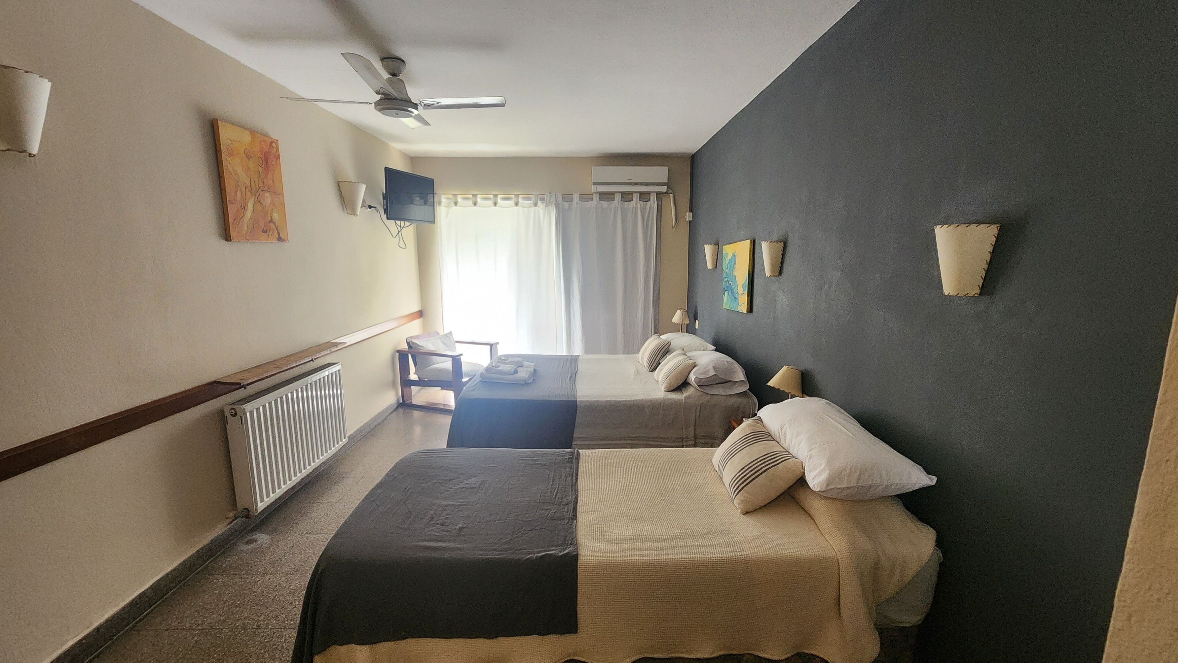 A hotel room with two beds, a ceiling fan, a radiator, a TV, and a window with curtains.