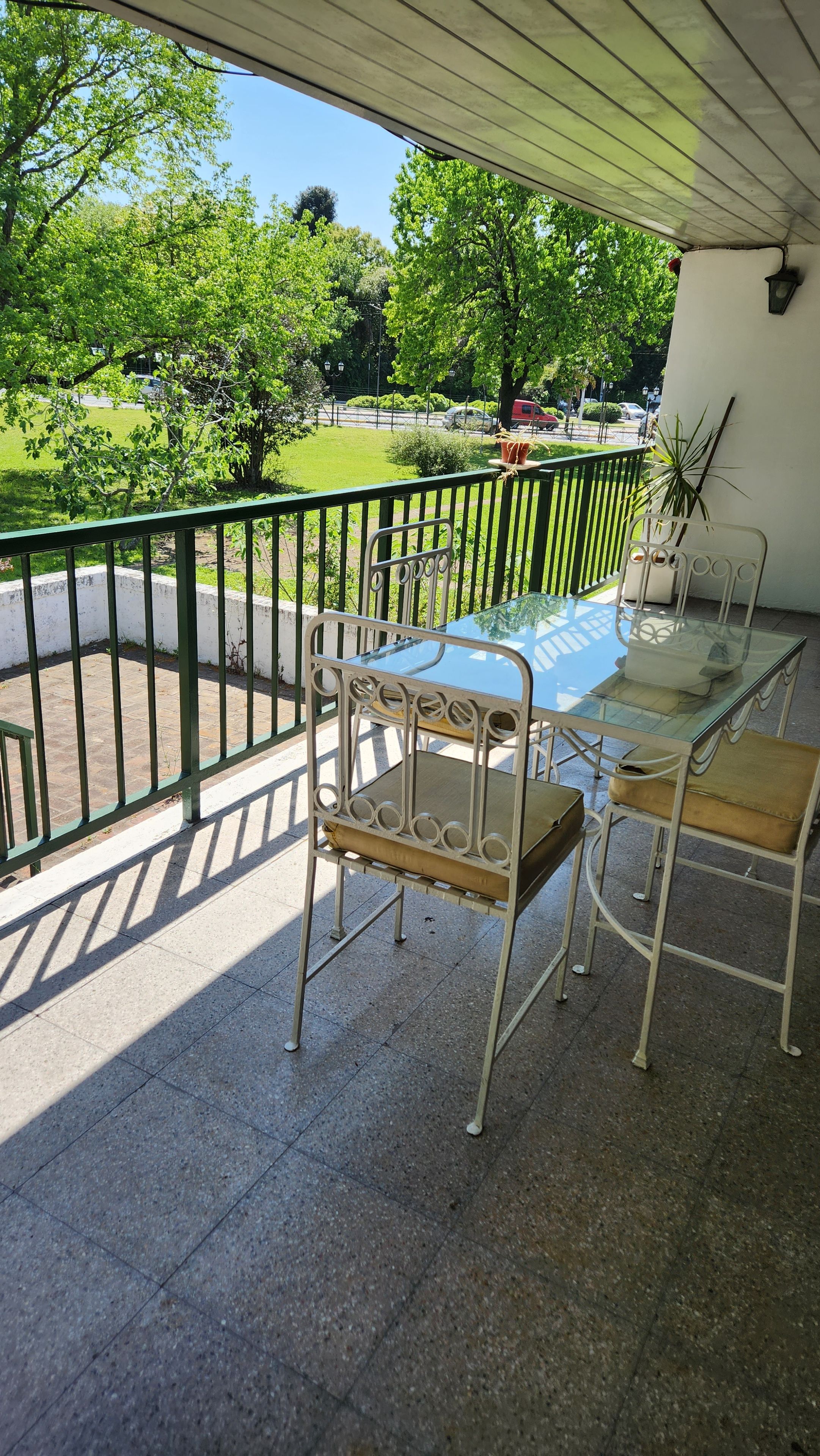 Un balcón con mesa y sillas de vidrio, una planta en maceta y vistas a un parque verde.