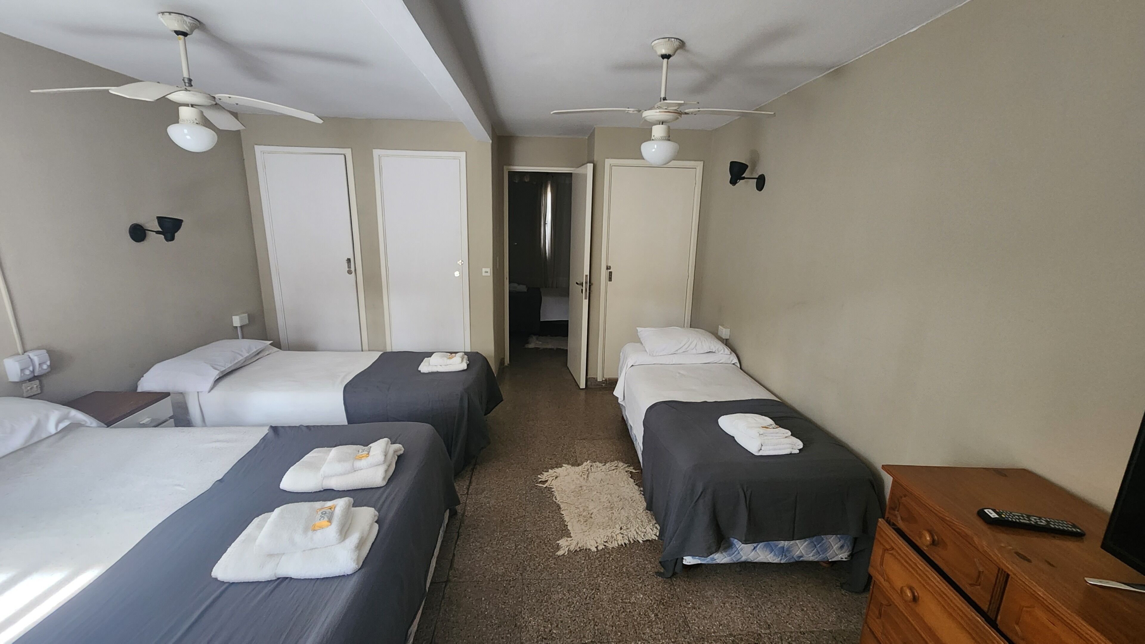 A hotel room with three beds, a wooden dresser, a desk with a computer, and a ceiling fan.
