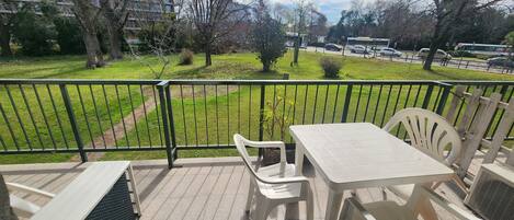 Un balcon avec une table et des chaises, une vue sur un espace verdoyant et une route où l’on aperçoit des véhicules au loin.