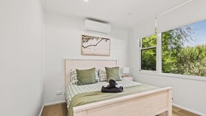 Ein Schlafzimmer mit Bett, einem Fenster mit Blick auf Grünflächen, einer Klimaanlage und einem an der Wand befestigten Kunstwerk.