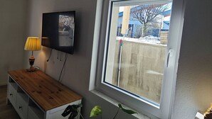 A room with a large window, a wooden cabinet, a television mounted on the wall, and a plant on the windowsill. - Charming Family Apartment in Wutöschingen With Garden View (Wutoeschingen)