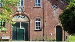 Un edificio de ladrillo con ventanas arqueadas y una puerta verde, rodeado de follaje.