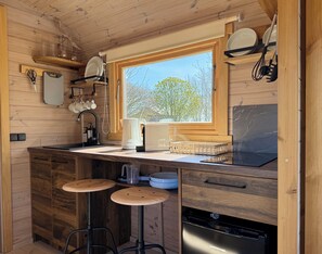 A compact kitchen with a window, wooden cabinets, a sink, stools, and a dishwasher. - Tiny House Säbelschnäbler Gut Osterhausen Butjadingen - Glamping and nature (Butjadingen)