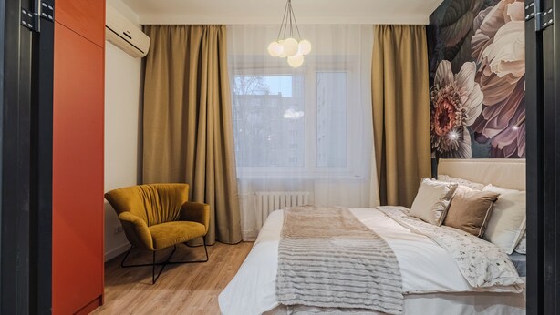 Ein Schlafzimmer mit einem Bett, einem Sessel, einem Fenster mit Vorhängen und einer Wand mit floralem Wandbild.