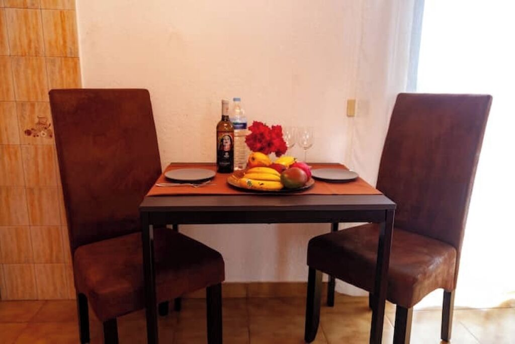 Una mesa de comedor con dos sillas, una botella de licor, un cuenco de fruta y un jarrón con flores.