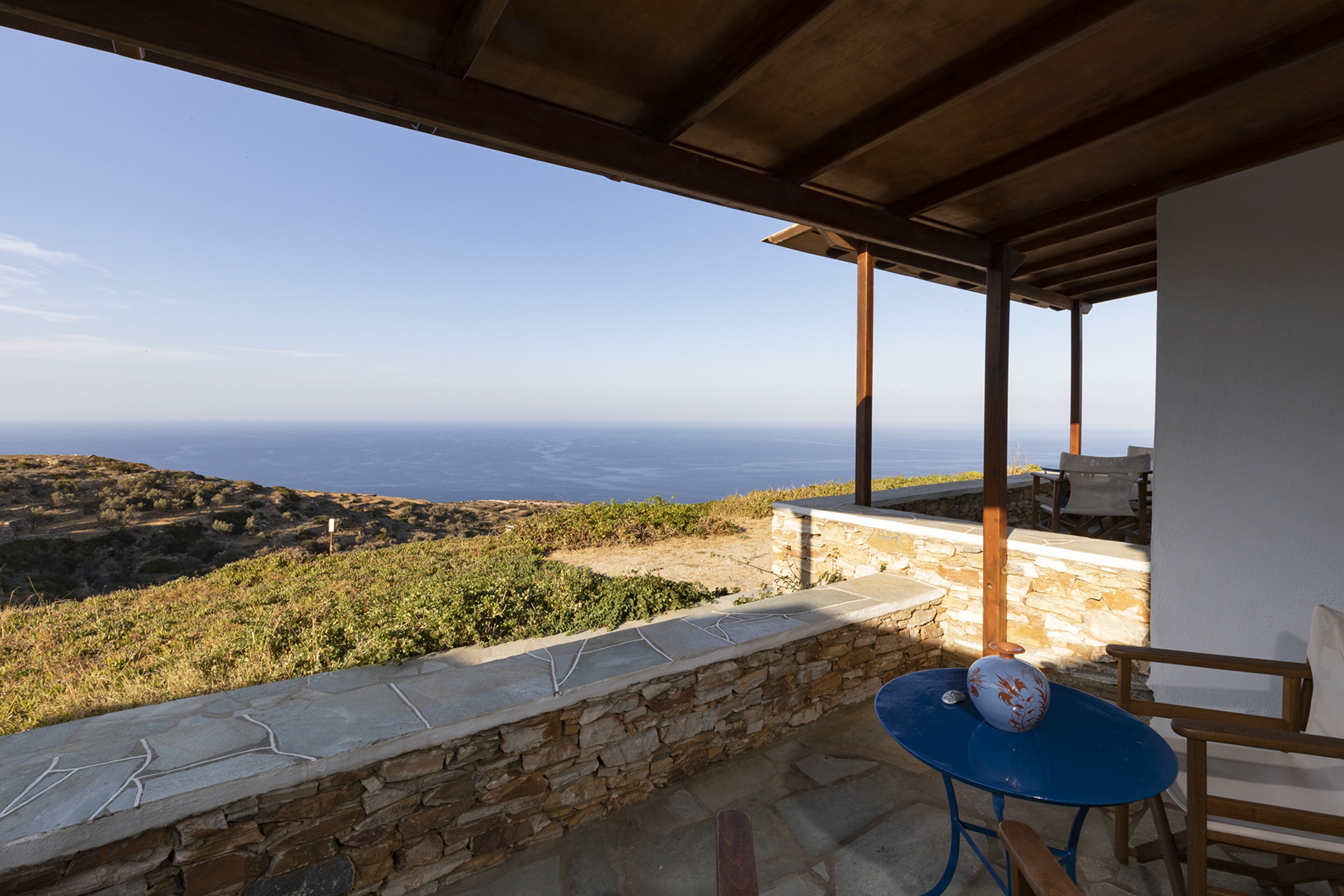 A terrace with a stone wall, a blue table, and a white chair, overlooking a coastal landscape.