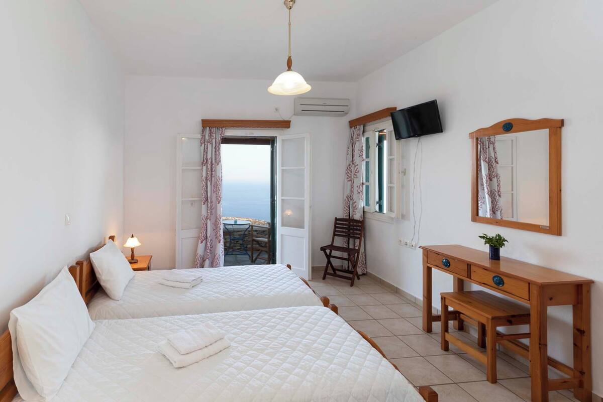 A hotel room with two beds, a wooden desk, a chair, a TV, and a balcony with a view.