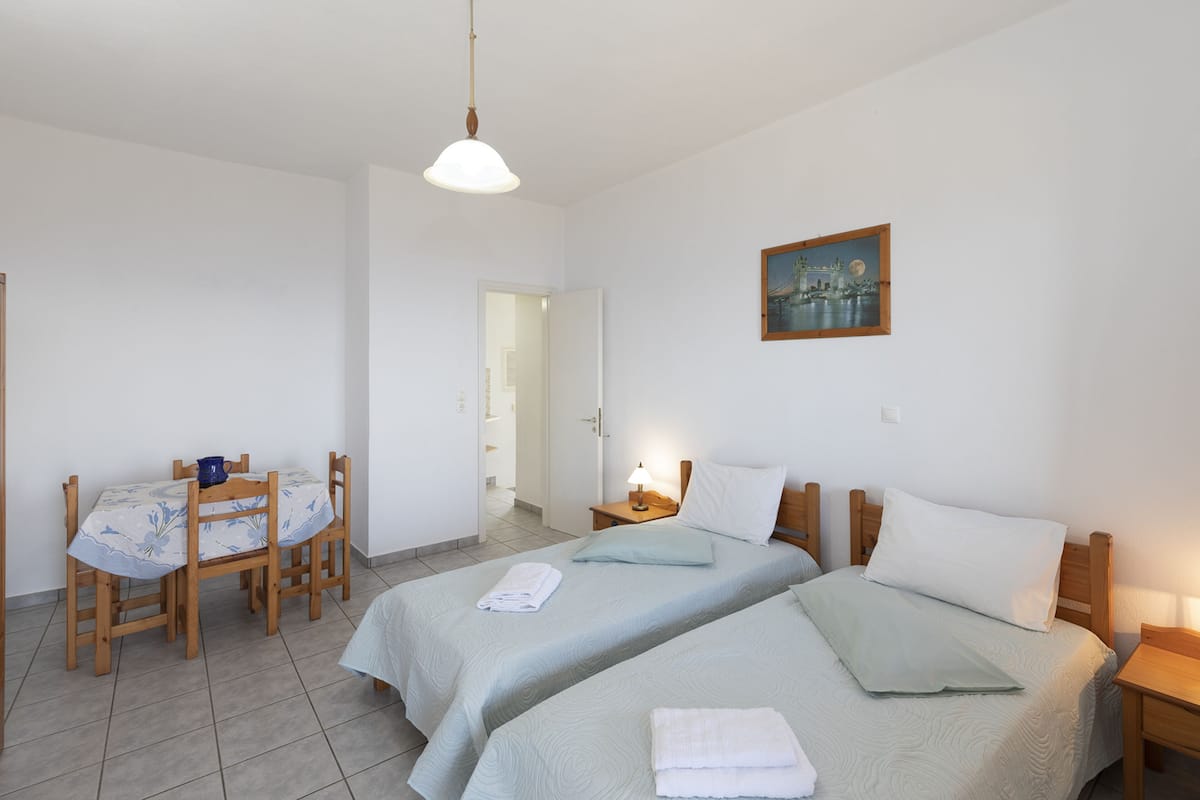 A room with two beds, a table with chairs, a painting on the wall, and a door leading to another room.