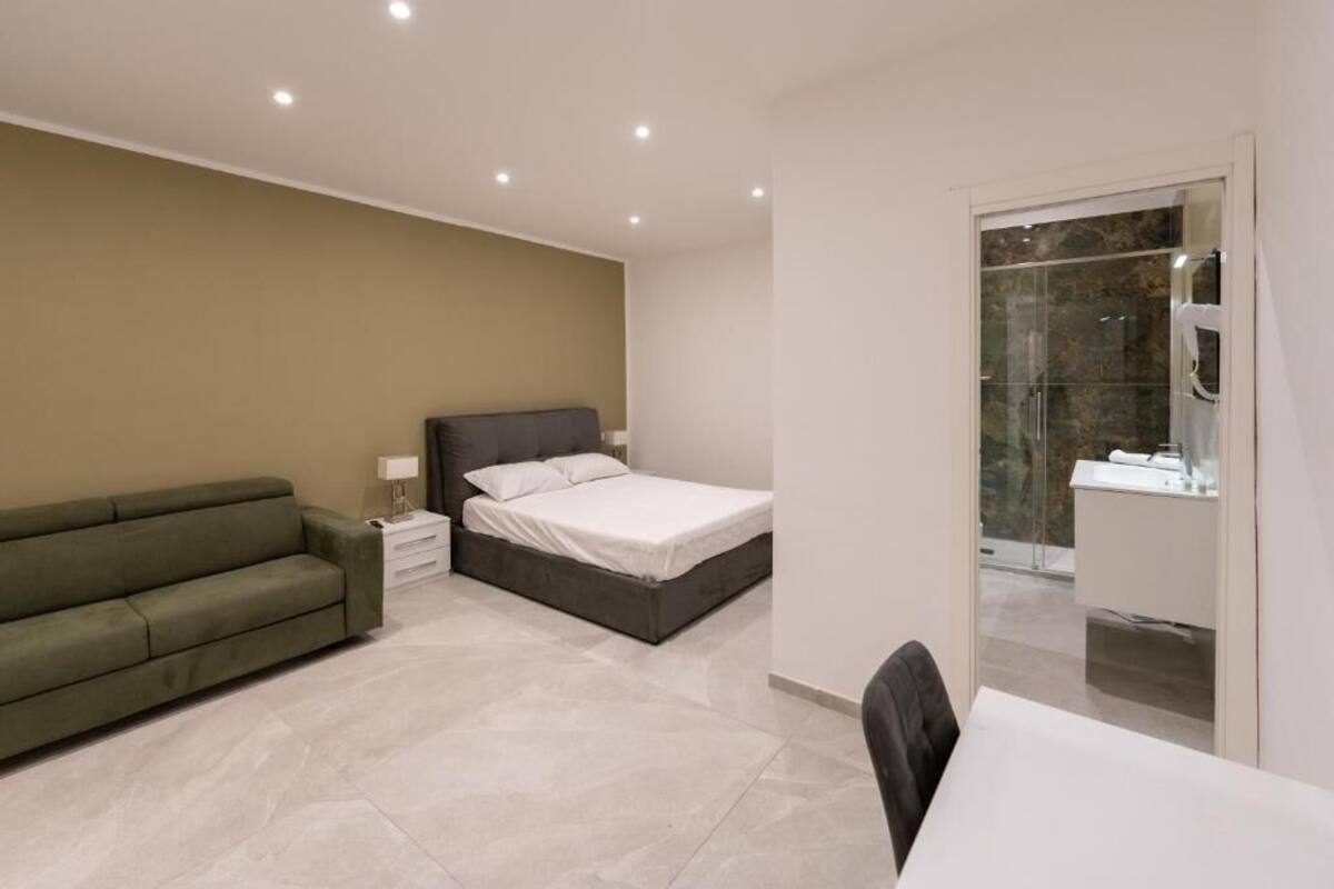 A modern bedroom with a bed, a green sofa, a bedside table, and a bathroom visible through a glass door.