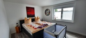 Une chambre à coucher avec un lit, un berceau, une fenêtre avec des stores, une horloge murale et un tableau.