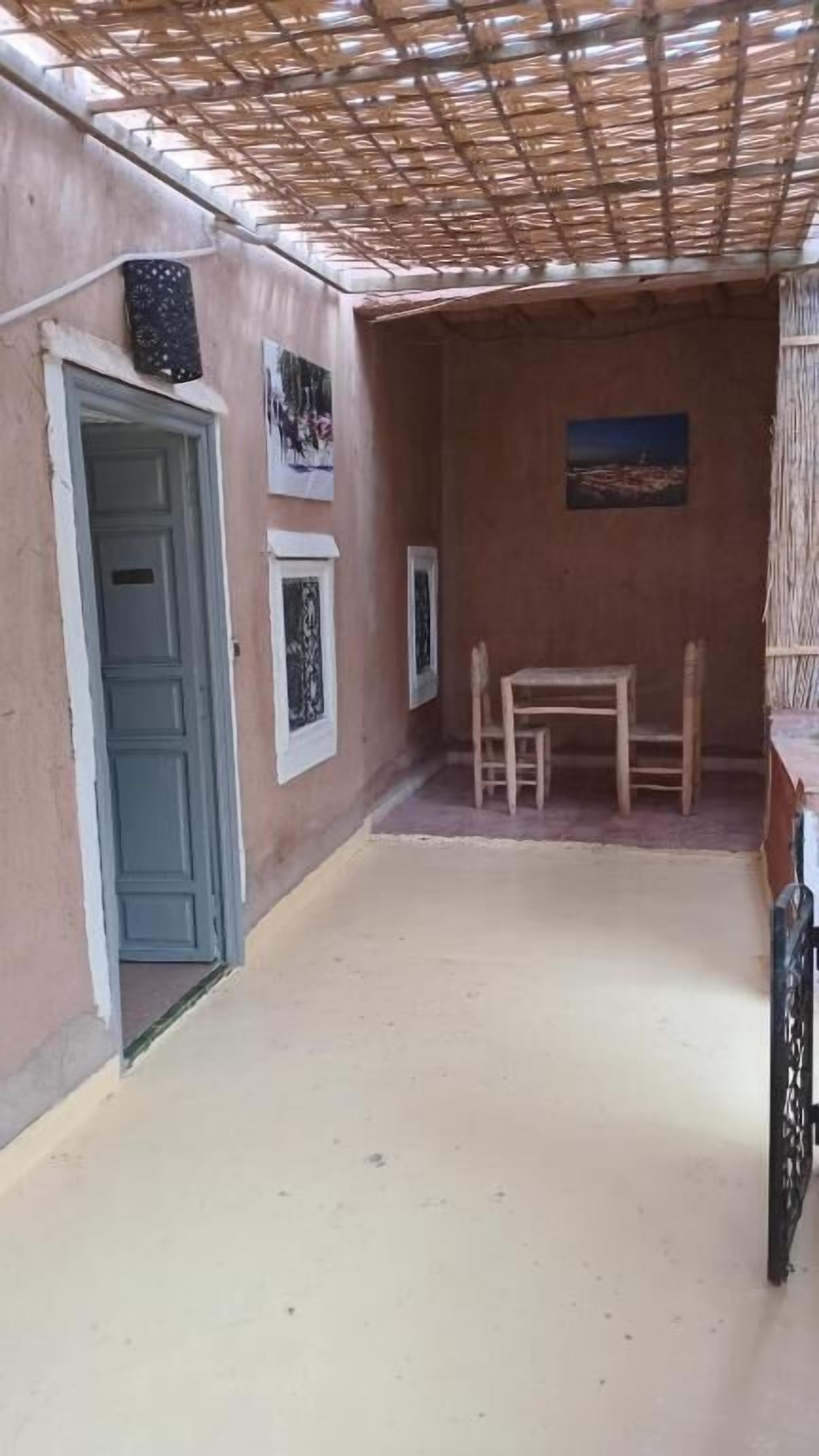 A covered outdoor area with a door, two framed pictures on the wall, a table with chairs, and a thatched roof.