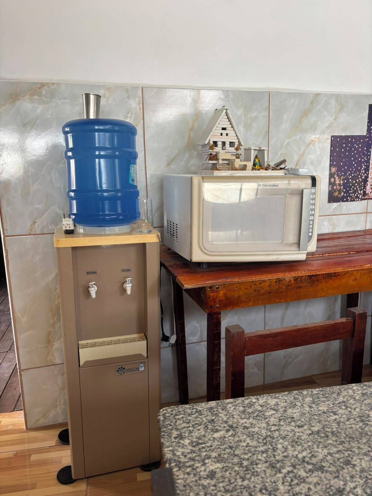 Uma kitchenette compacta com um dispensador de água, um micro-ondas e um pequeno modelo de casa sobre uma mesa de madeira.