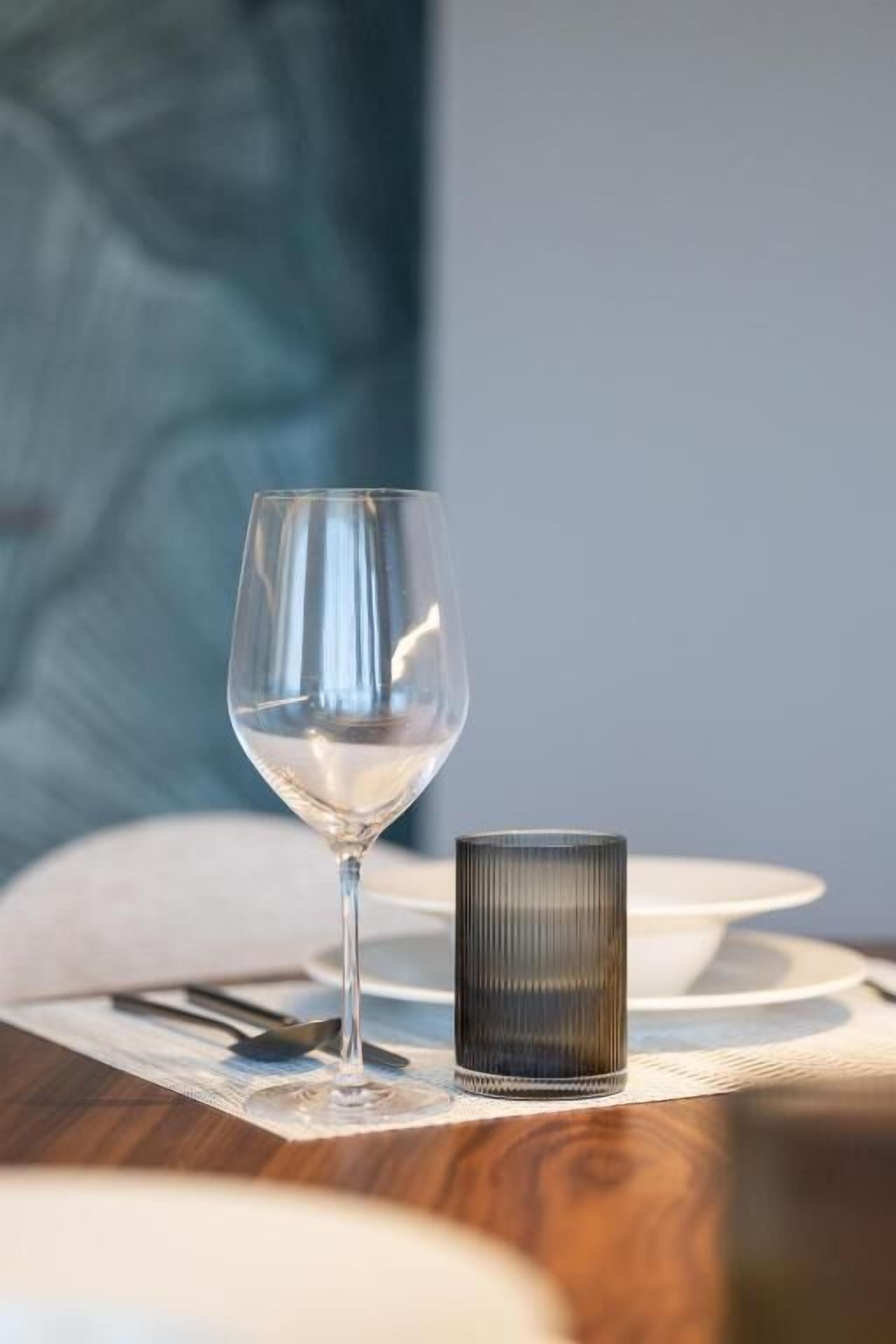 A clear glass wine glass and a ribbed candle holder on a wooden table with a placemat.