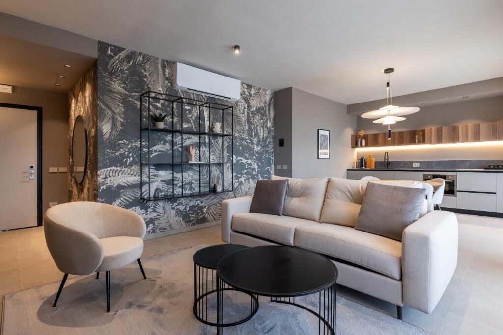 A modern living room with a sofa, two armchairs, and a coffee table. There is a kitchen in the background with a built-in oven and a pendant light.