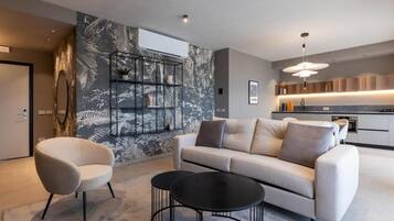 A modern living room with a sofa, two armchairs, and a coffee table. There is a kitchen in the background with a built-in oven and a pendant light.