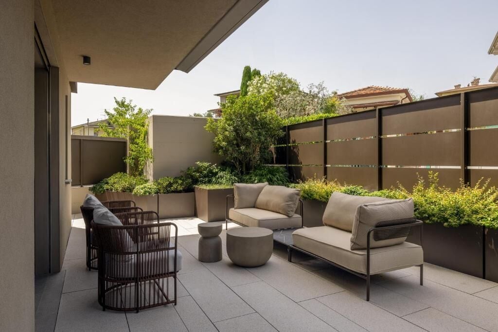 An outdoor patio area with modern furniture, including a sofa and chairs, surrounded by greenery and a metal fence.