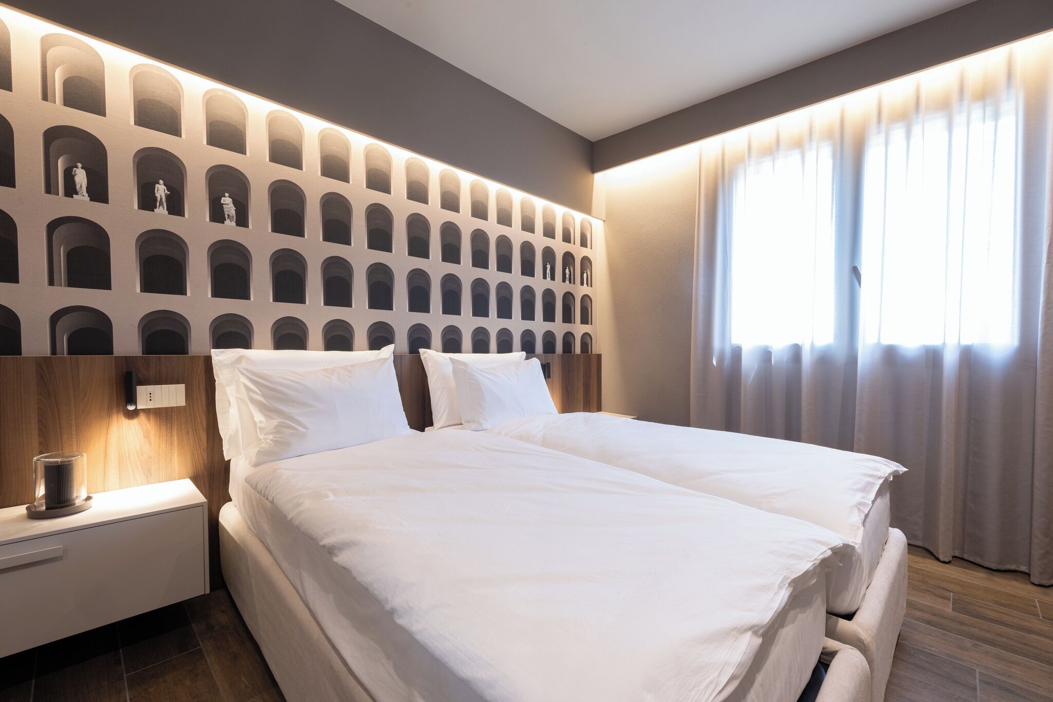 A modern hotel room with a large bed, a nightstand, a wall-mounted shelf with statues, and a large window with curtains.