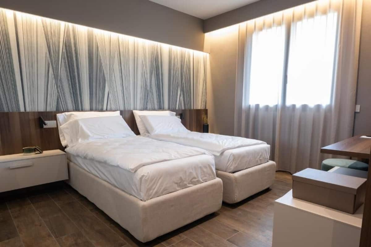 A modern hotel room with two beds, a bedside table, a desk, and a large window with curtains.