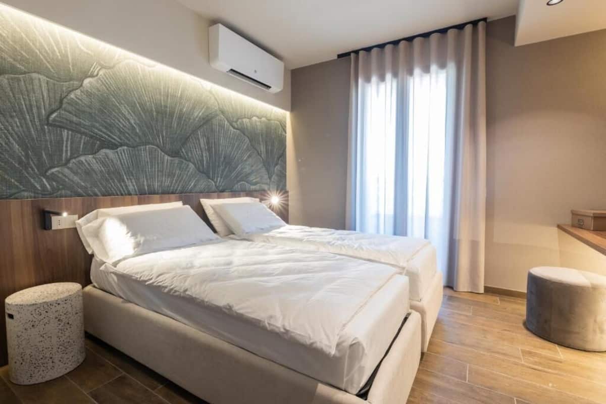 A modern hotel room with a large bed, a headboard with a textured design, a small round table, and a window with curtains.