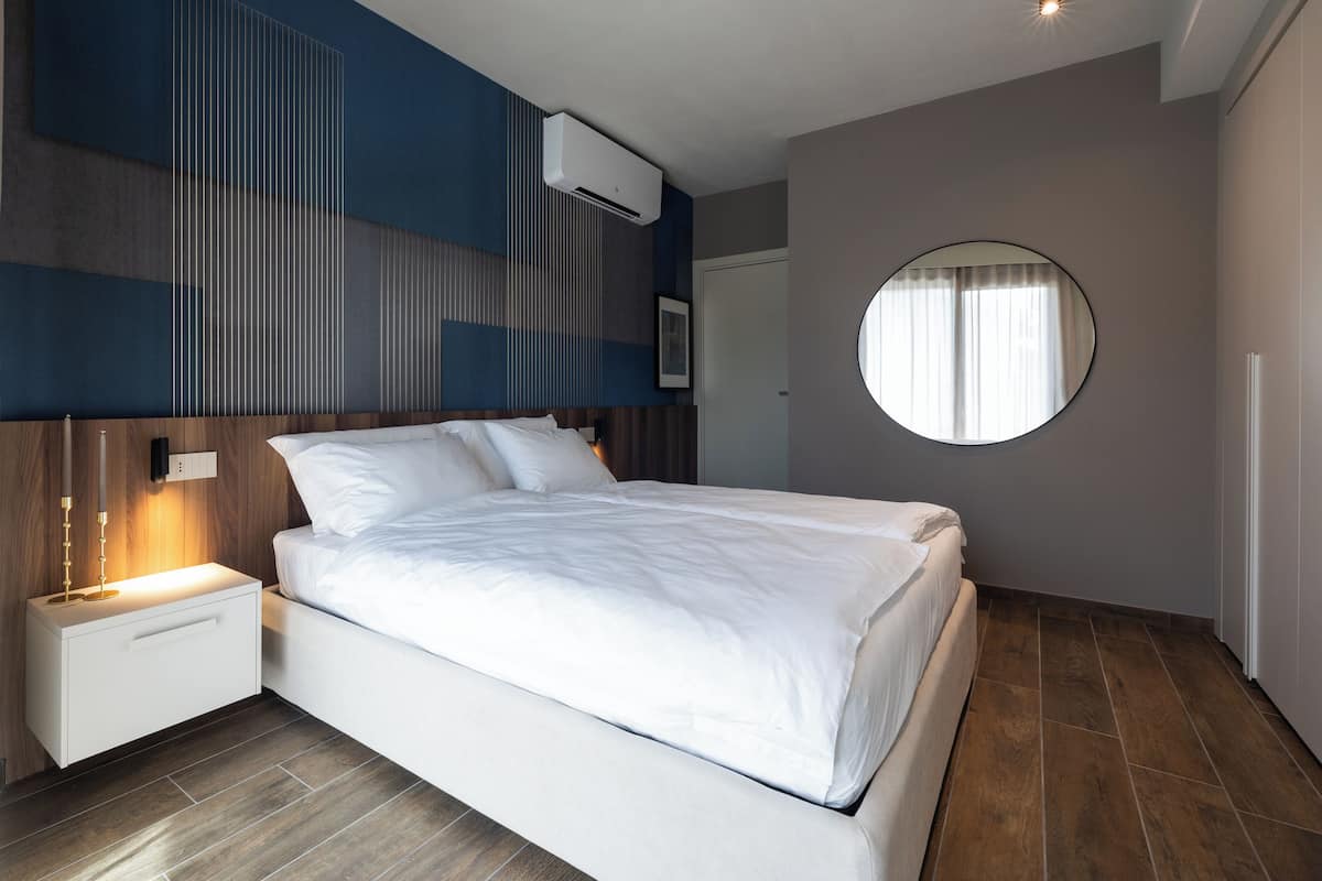 A modern bedroom with a large bed, a bedside table with a lamp, a round mirror, and an air conditioning unit on the wall.