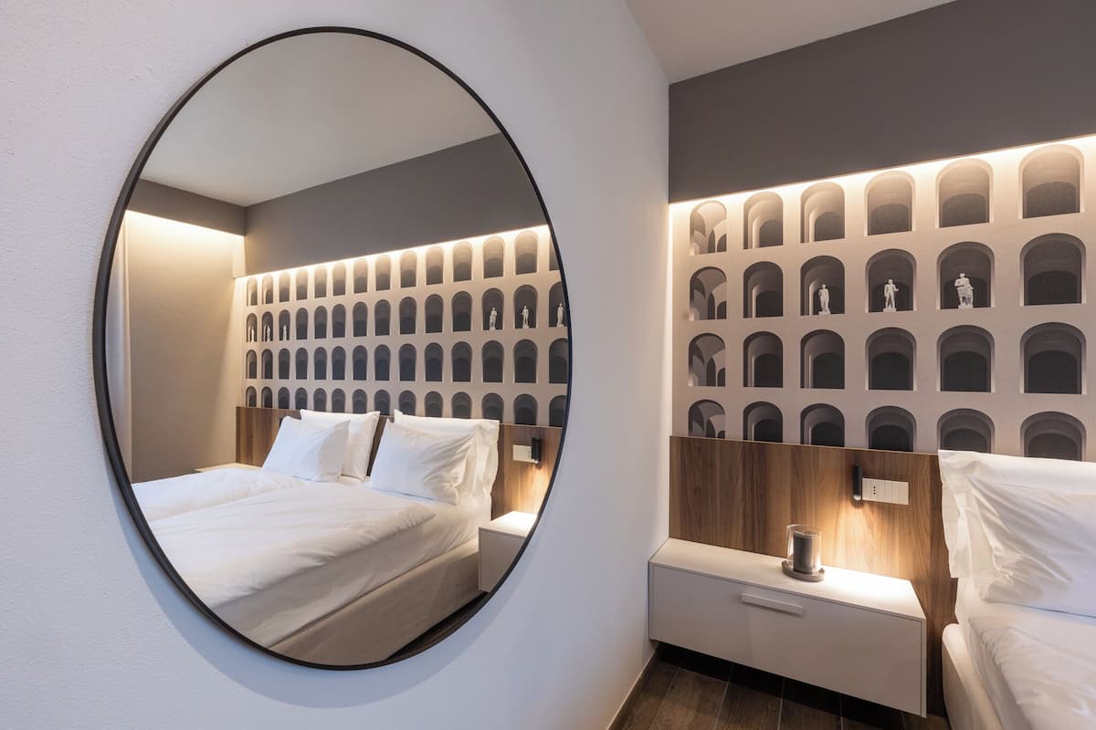 A modern hotel room with a circular mirror, a neatly made bed, a nightstand with a lamp, and a wall with a pattern of arches.