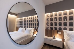 A modern hotel room with a circular mirror, a neatly made bed, a nightstand with a lamp, and a wall with a pattern of arches. - Stemor srl (Desenzano del Garda)