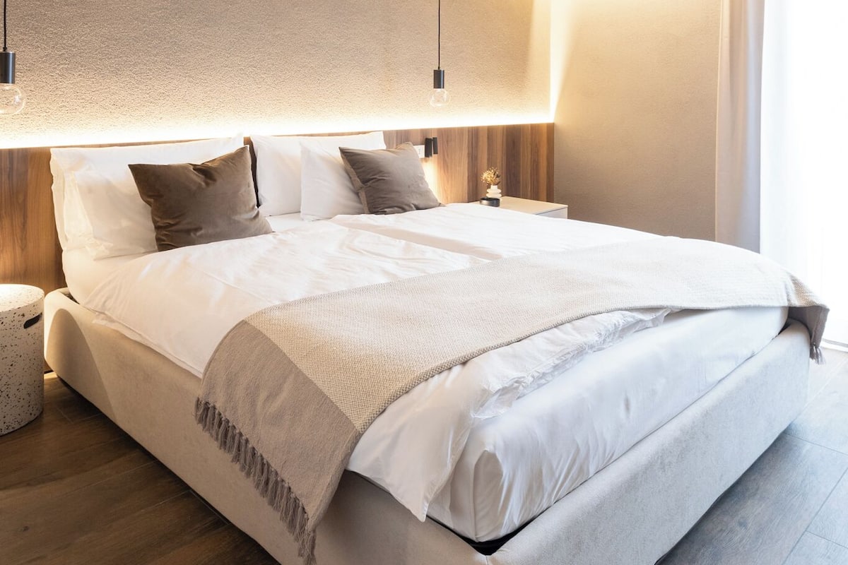 A modern bedroom with a large bed, wooden headboard, bedside tables, and a wall-mounted light fixture.