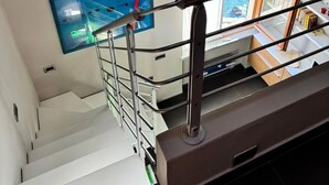 Eine Treppe mit Metallgeländern führt zu einer Zwischenebene. Ein Fenster mit Vorhängen ist sichtbar, und eine Uhr hängt an der Wand.