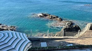 Eine Terrasse mit zwei Liegestühlen und einem gestreiften Sonnenschirm mit Blick auf eine felsige Küstenlinie und das Meer.