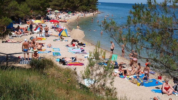 En overfyldt strand med folk, der solbader og svømmer, omgivet af træer og havet.