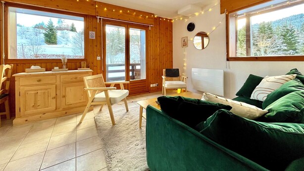 A cozy room with wooden walls, a green sofa, a dining table with chairs, and large windows with a view of trees. - Apartment with mountain and slope views (Combloux)