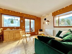 Una habitación acogedora con paredes de madera, un sofá verde, una mesa de comedor con sillas y ventanas grandes con vistas a árboles.