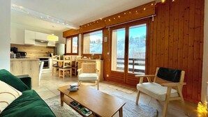 A cozy living room with a green sofa, a wooden coffee table, and a TV. There is a dining area with wooden chairs and a kitchen in the background. - Apartment with mountain and slope views (Combloux)