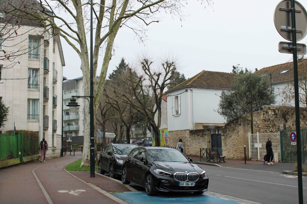 A street scene with a black car parked on the side, a pedestrian walking, and a building with a stone facade.
