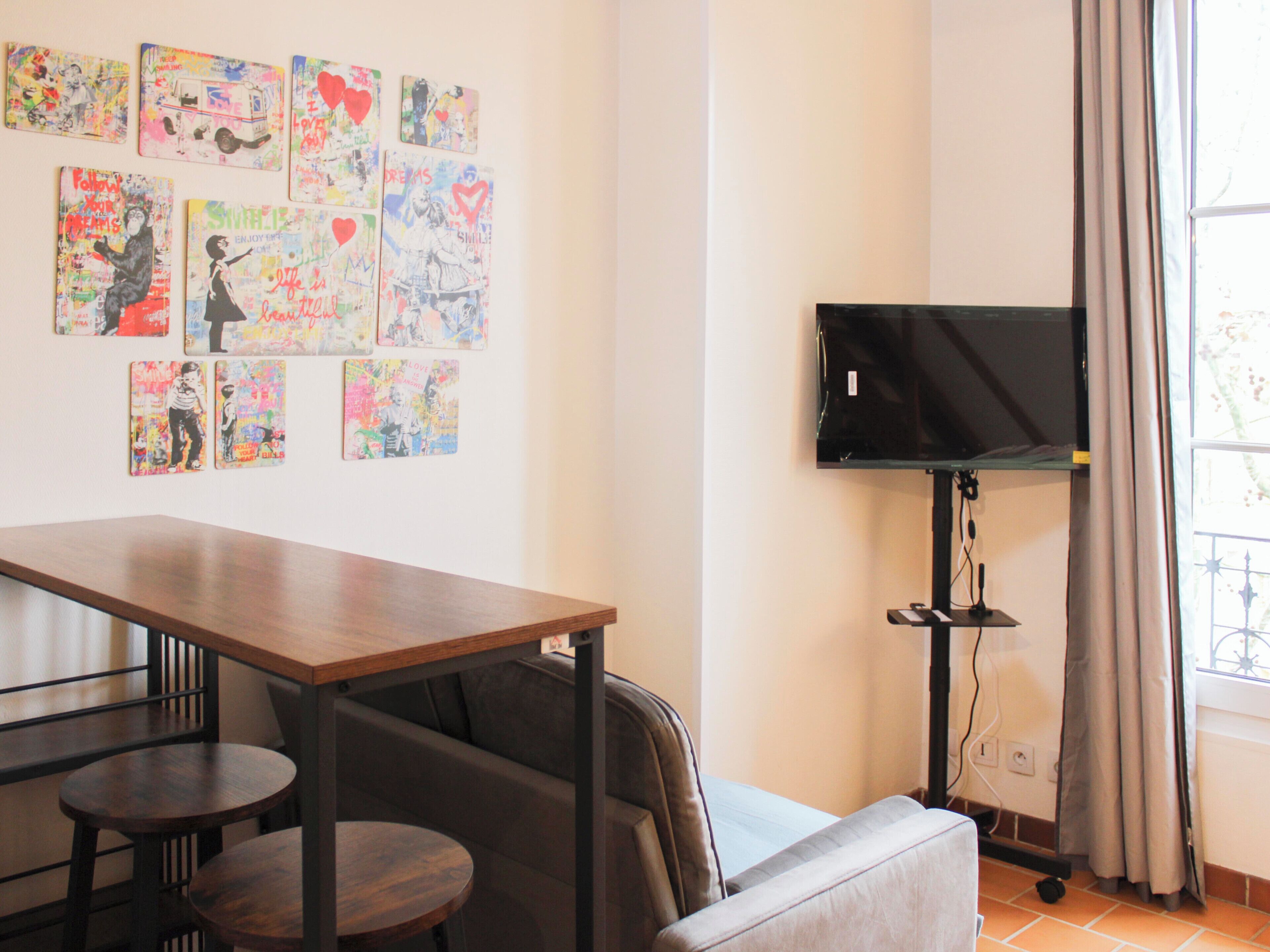 Une pièce comprenant une longue table, deux chaises, un canapé, un téléviseur à écran plat fixé sur un meuble, et une fenêtre avec des rideaux.