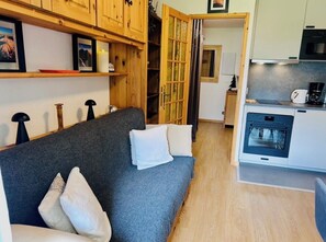 A cozy living area with a sofa, pillows, wooden shelves, a kitchen with a microwave, and a sliding door leading to another room. - Renovated flat with Mont Blanc's view | Parking (Chamonix-Mont-Blanc)