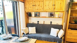 Een gezellige kamer met een grijze bank, houten kasten en een eethoek met een tafel gedekt voor twee personen.