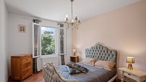 Une chambre à coucher avec un grand lit, une commode en bois, une table de chevet, un lustre et une fenêtre avec des rideaux.
