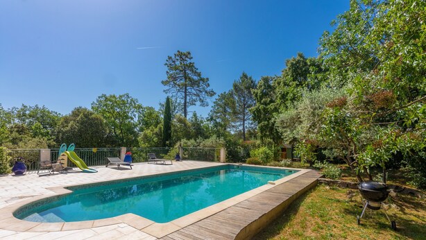 Une piscine rectangulaire avec une terrasse en bois, un toboggan et un barbecue, le tout installé dans un jardin.