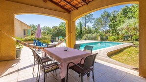 Une terrasse couverte avec une table et des chaises, un hamac et une vue sur une piscine et la verdure.
